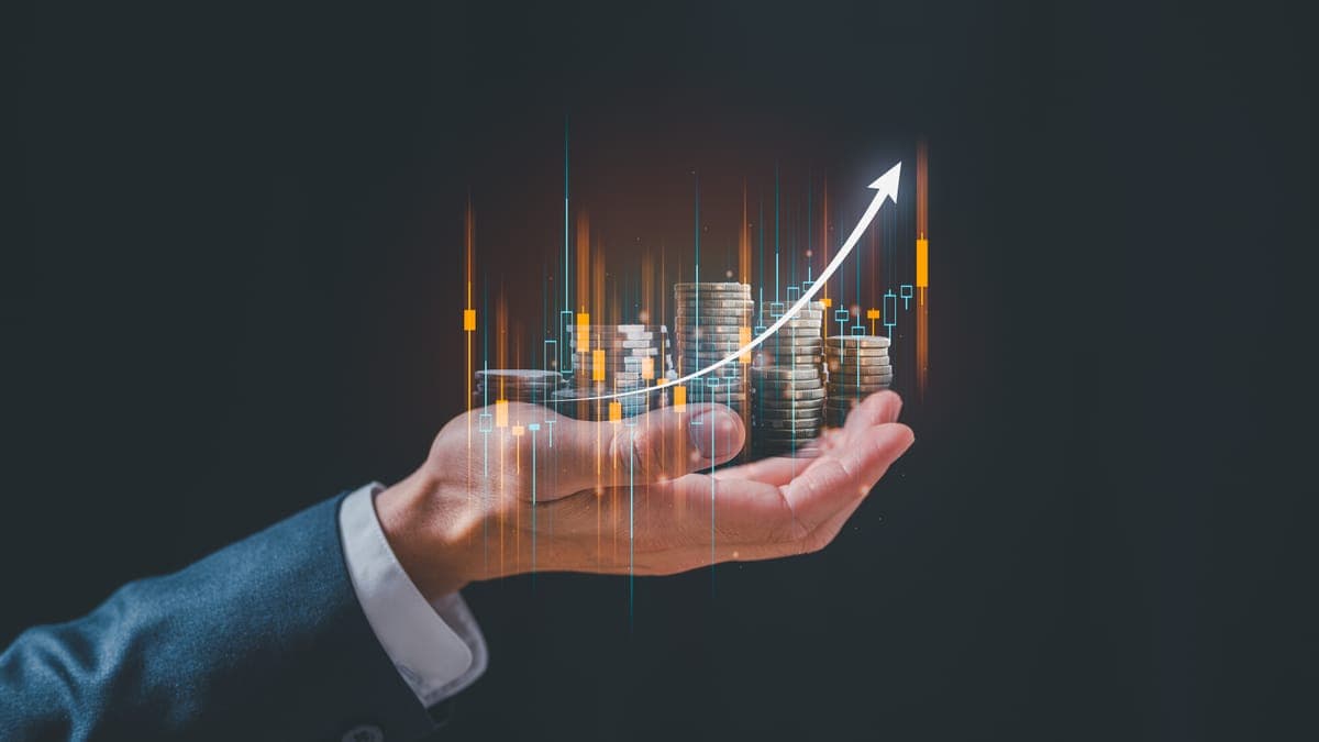 Hand im Anzug hält einen Münzstapel, darüber ein aufsteigender Kurspfeil mit Candlestick-Chart